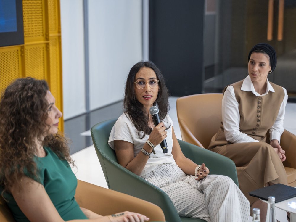 Karima Rahali-Sebti speaking on a panel, microphone in hand, alongside two other panellists